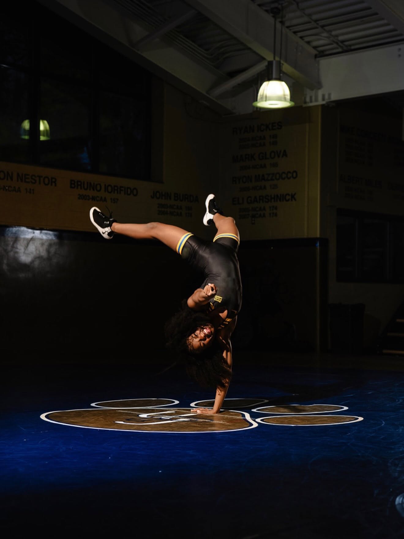Tamara Humphries holding a one-handed handstand on the mat under a single overhead lamp, upside down and laughing.